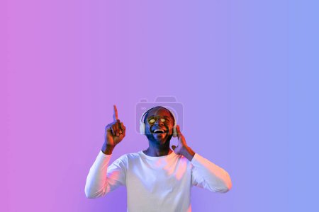 Téléchargez les photos : Beau et excité homme afro-américain d'âge moyen portant des lunettes de soleil à l'aide d'écouteurs sans fil modernes, pointant vers le haut de l'espace de copie pour la publicité et souriant, fond studio néon - en image libre de droit