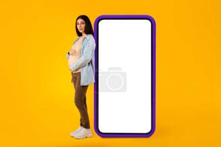 Foto de Mujer embarazada feliz apoyado en el teléfono inteligente grande con pantalla en blanco y abrazando su vientre, mujer expectante publicidad oferta en línea o aplicación de embarazo, longitud completa - Imagen libre de derechos