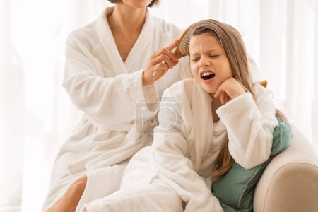 Foto de Mother combing tangled hair of her daughter after bath, little girl feeling pain from brushing, closeup shot of mom and female child wearing bathrobes having haircare routine at home, cropped - Imagen libre de derechos