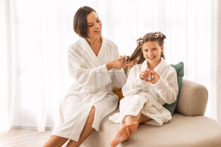 Photo for Mother And Daughters Wearing Bathrobes Making Hairstyles At Home, Caring Young Mom Winding Curlers To Her Cute Female Child While They Relaxing On Couch Together, Enjoying Beauty Day, Free Space - Royalty Free Image