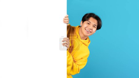 Photo for Happy Japanese Teen Boy Posing With Blank Paper Poster Smiling To Camera Over Blue Background In Studio. Great Offer Advertisement Banner Concept. Mockup, Panorama - Royalty Free Image