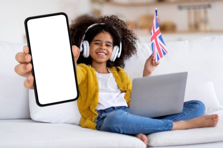 Téléchargez les photos : Smiling black preteen girl holding british flag and showing big blank smartphone at camera while sitting on couch at home, happy child recommending app for study english online, collage, mockup - en image libre de droit