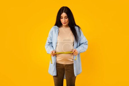 Téléchargez les photos : Jeune femme enceinte mesurant le ventre avec du ruban adhésif et le regardant, vérifiant la croissance, le développement et la santé, debout sur fond de studio jaune, espace de copie - en image libre de droit