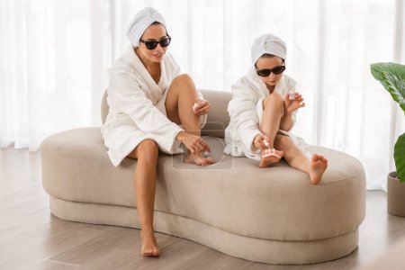 Téléchargez les photos : Little Girl And Her Mom Wearing Bathrobes And Sunglasses Doing Pedicure Together At Home, Happy Mother And Preteen Female Child Having Fun While Applying Nail Polish, Enjoying Self-Care Routine - en image libre de droit