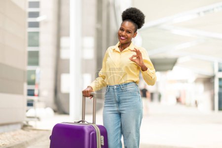 Foto de Trip Is Okay. Cheerful Black Tourist Woman Gesturing OK Posing With Suitcase Approving Tour Travel Offer Smiling To Camera Standing At Airport Outdoor. Vacation And Transportation Concept - Imagen libre de derechos