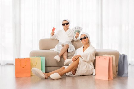 Foto de Mother And Daughter Wearing Bathrobes And Stylish Sunglasses Relaxing Among Bright Shopping Bags, Cool Family Holding Credit Card And Euro Cash Posing At Home, Enjoying Seasonal Sales And Purchases - Imagen libre de derechos