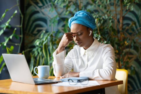Photo for Burnout at work. Tired upset millennial african american woman freelancer working from coffee shop, sitting at desk with closed eyes, touching nose bridge, using laptop and earpods, feeling exhausted - Royalty Free Image