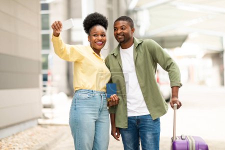 Téléchargez les photos : Des vacances. Époux afro-américains faisant Selfie au téléphone posant embrassant tenant passeport debout avec valise de voyage près de l'aérogare à l'extérieur. Couple familial voyageant à l'étranger - en image libre de droit