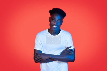 Téléchargez les photos : Cheerful happy cool young african american man in white t-shirt with bushy hair posing with arms crossed on chest in neon light over red studio background, black guy smiling at camera, copy space - en image libre de droit