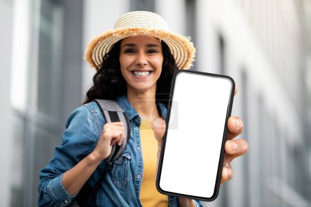 Téléchargez les photos : Happy young woman in straw hat holding smartphone with blank white screen while standing outdoors, smiling traveller woman showing empty cellphone, recommending mobile app or website, mockup - en image libre de droit