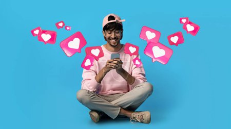 Téléchargez les photos : Glad young arabic guy in glasses has romantic chat with hearts on smartphone, enjoys message, sits on floor on blue studio background. Relationships and love remotely, app for dating, social networks - en image libre de droit