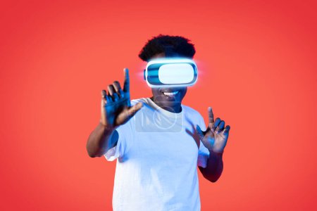 Téléchargez les photos : Happy cheerful young black guy in white t-shirt using modern VR goggles headset on red background in neon light, playing video games, virtual reality, simulators, trainers concept - en image libre de droit