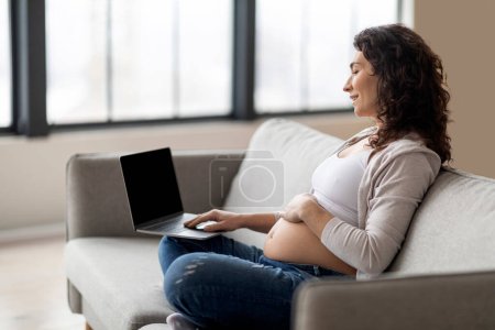 Téléchargez les photos : Femme enceinte souriante utilisant un ordinateur portable avec écran noir vierge tout en étant assis sur le canapé à la maison, belle jeune femme enceinte lecture grossesse Blog ou shopping en ligne sur ordinateur, Mockup - en image libre de droit