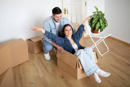 Photo for Cheerful Young Couple Riding Inside Cardboard Box While Having Fun On Moving Day, Happy Millennial Spouses Relocating To Their New Home, Fooling Together While Unpacking Things, Copy Space - Royalty Free Image