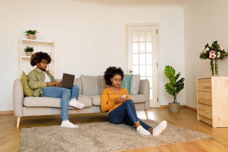 Téléchargez les photos : Black Couple Using Laptop And Mobile Phone Browsing Internet Sitting In Modern Living Room Indoors. Young Spouses Posing With Gadgets At Home On Weekend. Technology And Social Media Concept - en image libre de droit