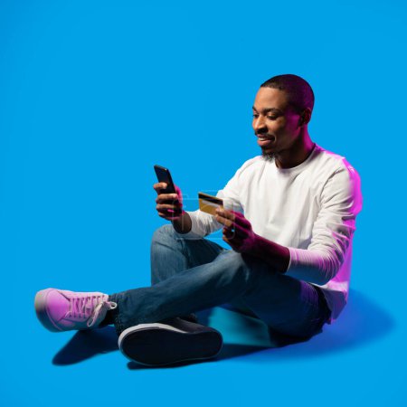 Téléchargez les photos : Cheerful handsome young black man sitting on floor on blue background in neon light, using smartphone and holding credit card, online banking concept, e-commerce, copy space - en image libre de droit