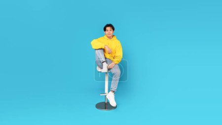 Téléchargez les photos : Korean Teenager Boy Sitting On Bar Stool Looking At Camera Posing Over Blue Studio Background, Wearing Jeans And Yellow Hoodie. Full Length Shot Of Asian Student Guy. Panorama - en image libre de droit