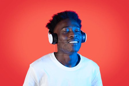 Téléchargez les photos : Joyful peaceful young black guy in white t-shirt listening to music with closed eyes and smiling, using modern wireless headphones in neon light, red background, closeup, copy space - en image libre de droit