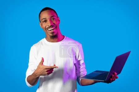 Téléchargez les photos : Joyeux joyeux beau jeune homme noir pointant vers ordinateur portable PC moderne dans sa main et souriant à la caméra, travaillant en ligne sur fond de studio bleu dans la lumière au néon, l'espace de copie. Concept d'emploi à distance - en image libre de droit