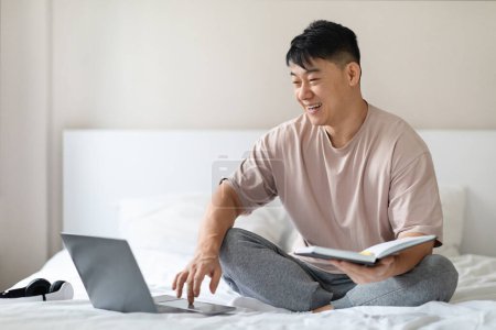 Téléchargez les photos : Good morning. Relaxed handsome mature asian man independent contractor checking his agenda on notepad, using brand new laptop and smiling, staying in bed at home, wearing pajamas, copy space - en image libre de droit