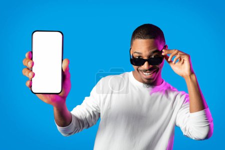 Foto de Cool handsome cheerful young black man in white showing modern cell phone with white empty screen, wearing sunglasses, recommending nice app in neon light on blue background, mockup - Imagen libre de derechos