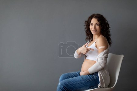 Foto de Hermosa mujer embarazada sentada en la silla y mostrando el brazo después de la vacunación contra el Coronavirus, sonriendo embarazada señalando el hombro con vendaje adhesivo mientras posa sobre fondo gris - Imagen libre de derechos