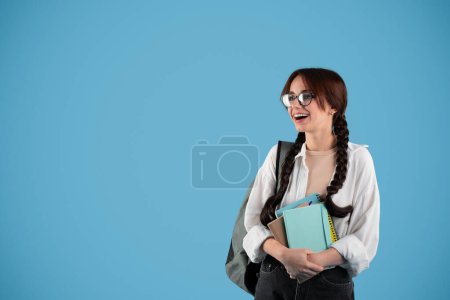 Sorprendido adolescente feliz hembra europea inteligente con coletas en gafas con mochila sostiene cuadernos y mirando el espacio vacío aislado sobre fondo de estudio azul. Anuncio y oferta de estudio y conocimiento