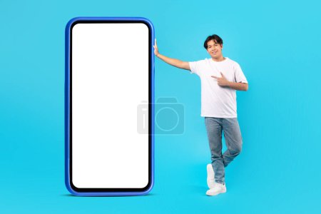 Photo for Happy Korean Teen Boy Pointing At Large Smartphone Empty Screen Recommending Application Standing Near Gadget Over Blue Studio Background. Great Mobile Offer. Mockup - Royalty Free Image