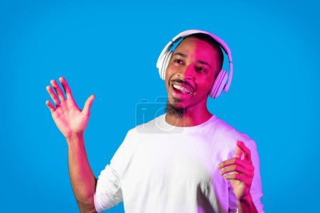 Téléchargez les photos : Happy cheerful handsome talented young african american guy singing songs and moving hands, using wireless headphones on blue studio background in neon light, copy space. Hobby concept - en image libre de droit