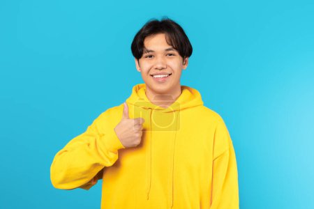 Téléchargez les photos : I Like It. Happy Asian Teenager Guy Gesturing Thumbs Up In Approval Posing Smiling To Camera On Blue Studio Background. Portrait Of Teen Boy Approving Great Offer Wearing Yellow Hoodie - en image libre de droit