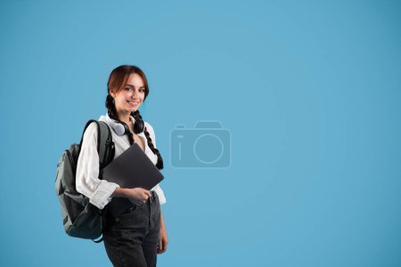 Téléchargez les photos : Happy teen european girl student with pigtails, backpack and headphones hold laptop, isolated on blue studio background. Gadget for learn and knowledge at school, tutoring, ad and offer, new normal - en image libre de droit