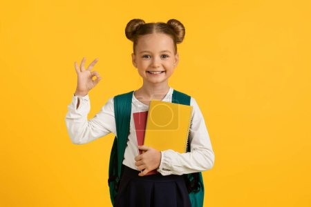 Téléchargez les photos : Cute Little Schoolgirl With Workbook In Hands Showing Ok Gesture, Happy Female Pupil With Backpack Gesturing Okay, Recommending Education Program While Standing Isolated Over Yellow Background - en image libre de droit