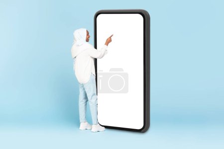 Photo for Muslim woman touching huge display panel of smartphone with blank white screen, standing on blue background, mockup, full body length - Royalty Free Image