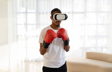 Photo for Young black guy fighter in VR headset and red boxing gloves having virtual boxing workout at home. African American man training via augmented reality experience, copy space - Royalty Free Image