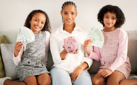 Photo for Cheerful Black Mom And Daughters Holding Piggybank With Family Savings And Paper Money Smiling To Camera Sitting On Couch At Home. Selective Focus On Piggy Bank - Royalty Free Image