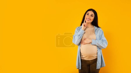 Téléchargez les photos : Heureuse femme enceinte coûteuse choisissant le nom pour bébé, posant sur fond jaune, panorama avec espace libre. Rêver et imaginer la vie maternelle - en image libre de droit