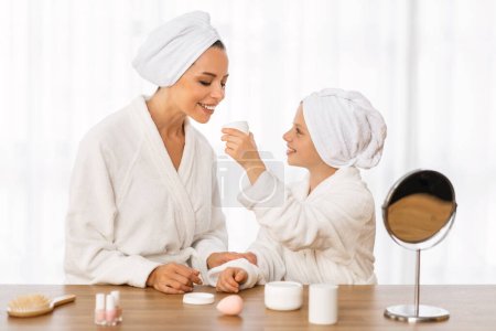 Photo for Mother And Little Daughter In Bathrobes Testing New Cosmetics While Making Beauty Routine At Home, Happy Young Mom And Female Child Checking Jar With Moistursing Cream, Enjoying Skincare Products - Royalty Free Image