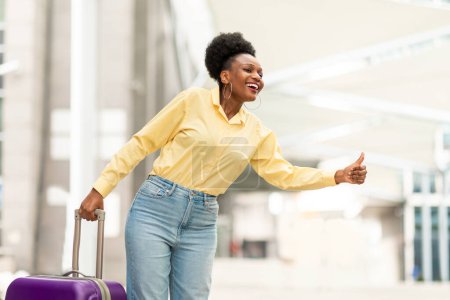 Téléchargez les photos : Joyeux touriste noire attrapant un taxi avec geste de la main regardant à côté debout avec une valise de voyage à l'aéroport en plein air. Tourisme et Transport Concept - en image libre de droit