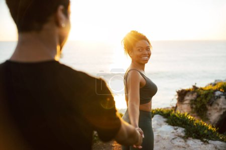 Foto de Feliz joven afroamericana femenina en ropa deportiva lleva a hombre a mano en la mañana en las rocas cerca del océano, disfrutar del ejercicio y el amanecer, al aire libre. Relaciones, amor, cuidado de la salud, entrenamiento y estilo de vida activo - Imagen libre de derechos