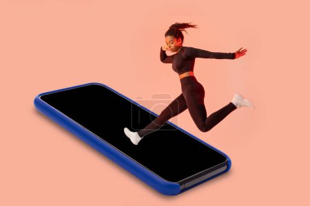 Photo for Fitness app. Black woman jumping on smartphone blank screen, having workout on peach studio background, mockup. Sport and technology concept - Royalty Free Image