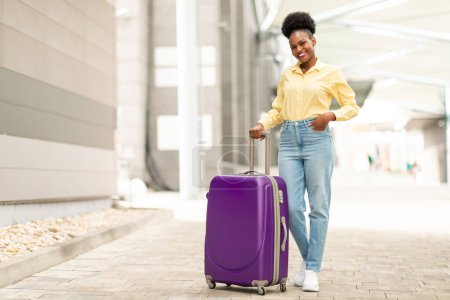 Téléchargez les photos : Femme afro-américaine joyeuse posant avec une valise de voyage souriant à la caméra debout à l'aéroport en plein air. Touriste Femme Publicité Vacances Tour Offre. Concept touristique. Longueur totale - en image libre de droit