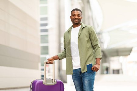 Téléchargez les photos : Joyeux voyageur noir homme debout avec une valise de voyage tenant passeport souriant à la caméra posant debout à l'aéroport. Tourné de heureux touristes masculins. Concept vacances et transport - en image libre de droit