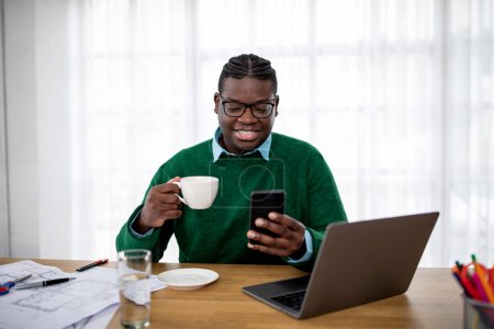 Foto de Toothy chico afroamericano usando Smartphone trabajando en línea en el ordenador portátil y tener café sentado en el escritorio en la oficina moderna. Carrera exitosa y concepto de estilo de vida empresarial - Imagen libre de derechos