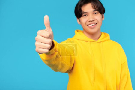 Photo for I Like It. Smiling Korean Teenager Boy Gesturing Thumbs Up To Camera, Approving Offer Posing Standing On Blue Background, Studio Shot. Selective Focus On Guys Hand. Approval Concept - Royalty Free Image