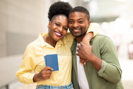 Téléchargez les photos : Voyage de vacances. Joyeux couple de touristes noirs posant embrassant et tenant des passeports souriant à la caméra debout à l'extérieur près du terminal de l'aéroport moderne. Offre de billets d'avion pas chers - en image libre de droit