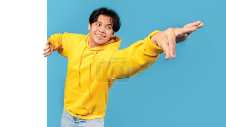 Photo for Smiling Korean Teenager Guy Posing With Blank Paper Board Holding And Showing Something To Camera On Blue Studio Background. Product Advertisement Banner. Panorama, Mockup - Royalty Free Image