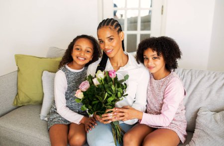 Photo for Mothers Day. Cheerful Black Girls Hugging Their Mommy Giving Her Bouquet Of Fresh Flowers Posing Sitting On Couch At Home. Family Of Three Smiling To Camera Celebrating Holiday Together - Royalty Free Image
