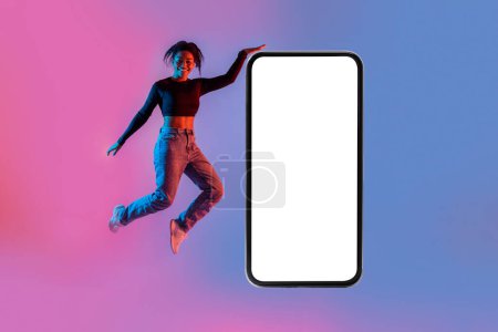 Photo for Cellphone screen mockup. Happy black woman jumping near giant cellphone with white blank screen, promoting new app, website or great offer in neon light, copy space, mockup - Royalty Free Image