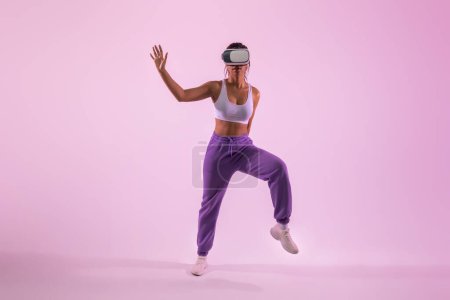 Foto de Ajuste señora afroamericana bailando y divirtiéndose en el metaverso mientras usa gafas de realidad virtual, explorando la tecnología 3D sobre fondo de luz de neón de estudio rosa - Imagen libre de derechos