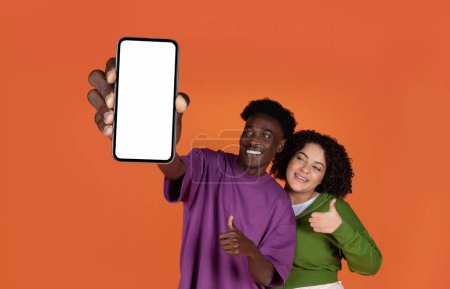 Téléchargez les photos : Mignon joyeux multiracial jeune couple hipsters montrant tout nouveau téléphone portable avec écran blanc et pouce vers le haut à la caméra, recommander belle sortir ensemble app, isolé sur fond rouge, maquette - en image libre de droit
